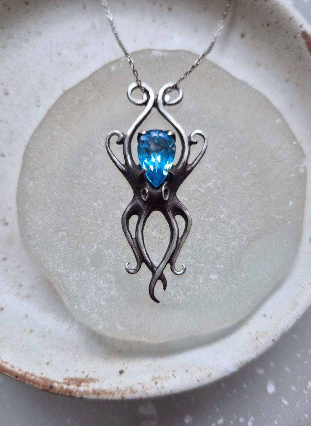 Close-up of silver Octopus necklace with faceted blue topaz, placed on frosted glass and ceramic dish