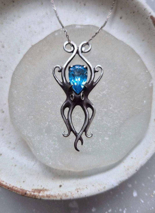 Close-up of silver Octopus necklace with faceted blue topaz, placed on frosted glass and ceramic dish