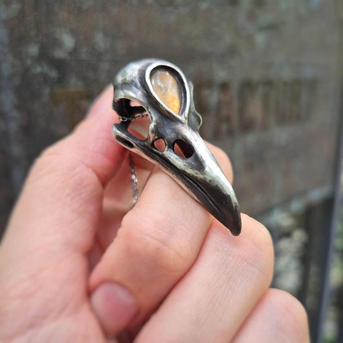 Golden labradorite raven skull necklace held in hand, showing cabochon glow and oxidized silver detail.
