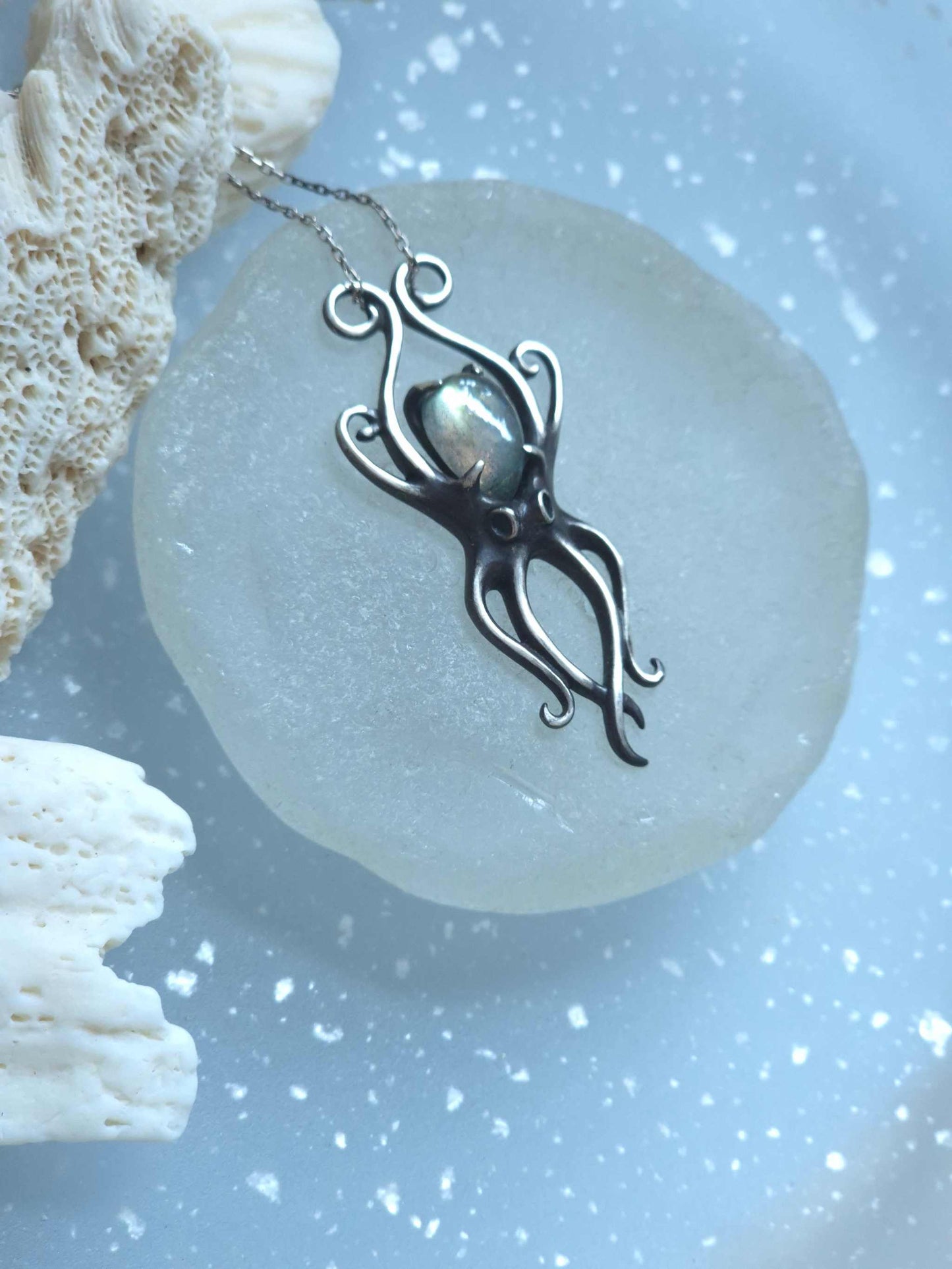 Flatlay of silver Octopus pendant resting on matte glass surface, surrounded by natural white coral
