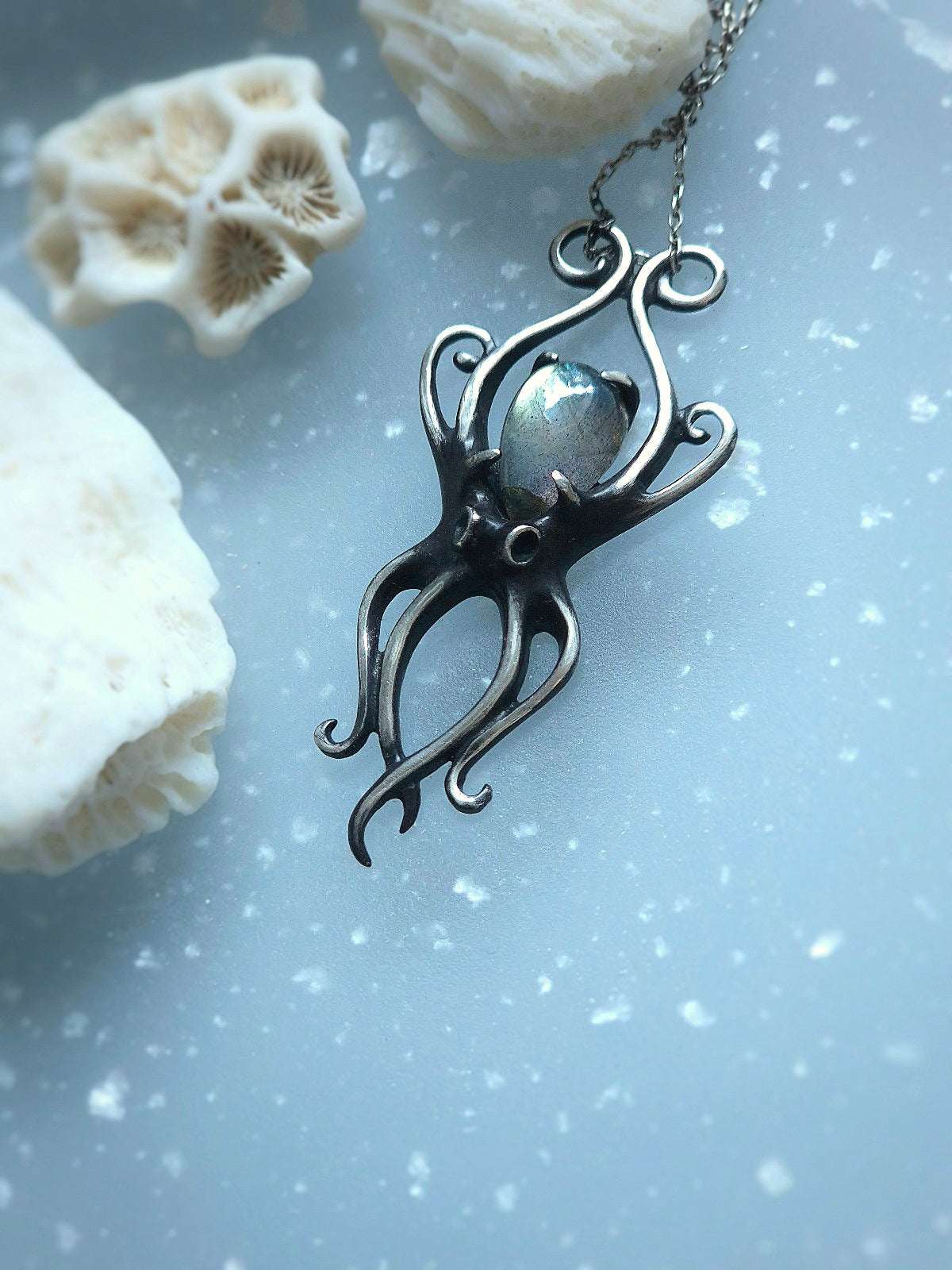 Flatlay of oxidized silver octopus pendant with gemstone, placed on blue background and white coral