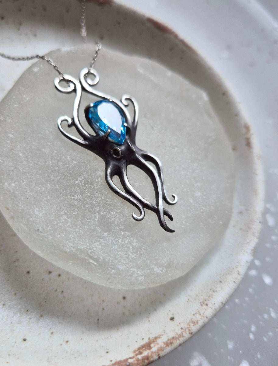 Angled view of silver Octopus necklace with vibrant ocean-blue gem on matte glass surface