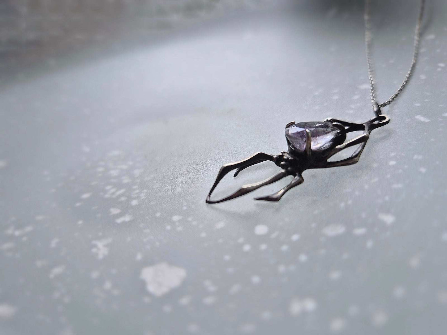 Side view of a silver spider pendant with quartz gem, on a glass surface with soft speckled light patterns