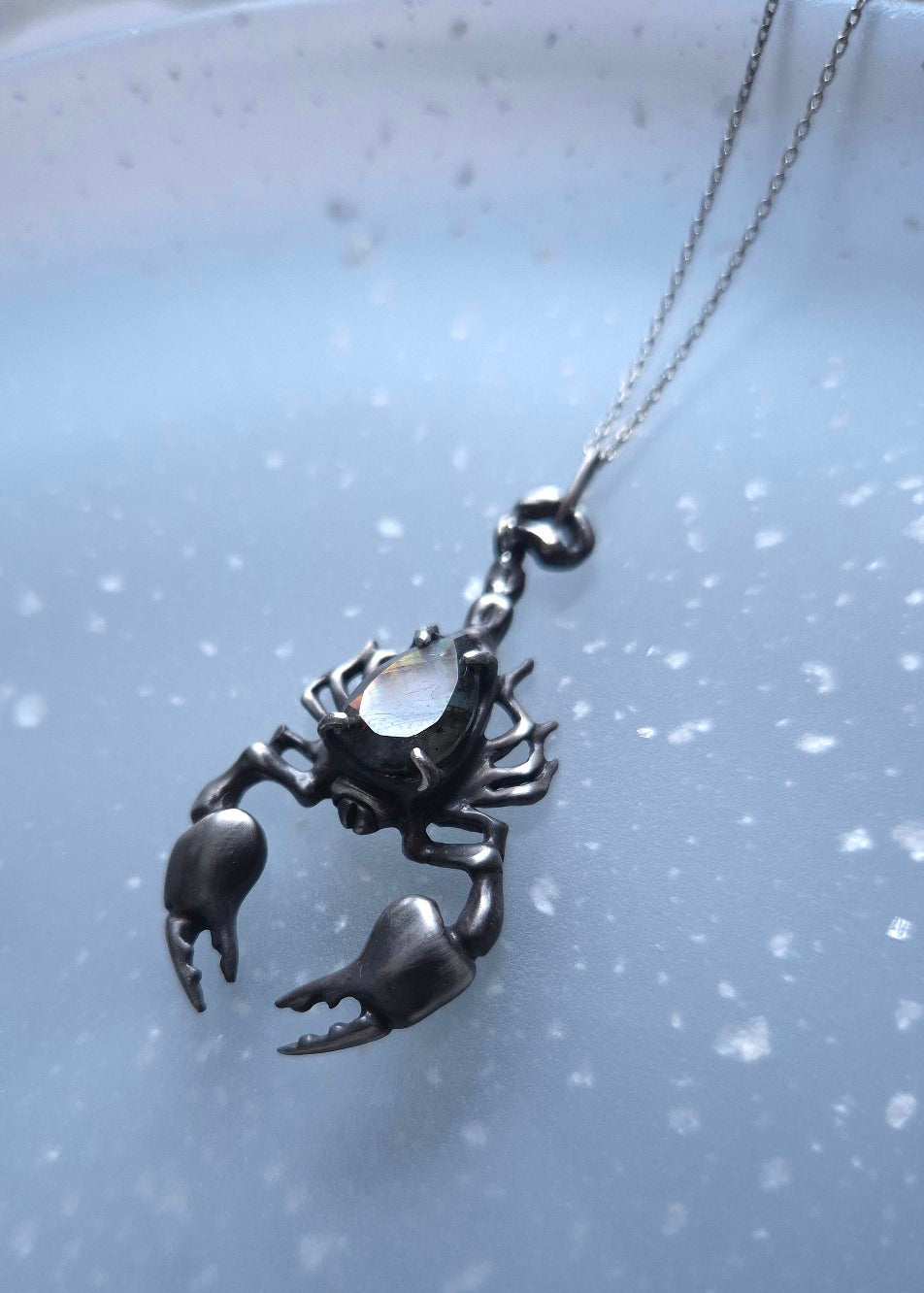 Dark silver Scorpion necklace with a teardrop labradorite with subtle light reflections, on a speckled blue surface
