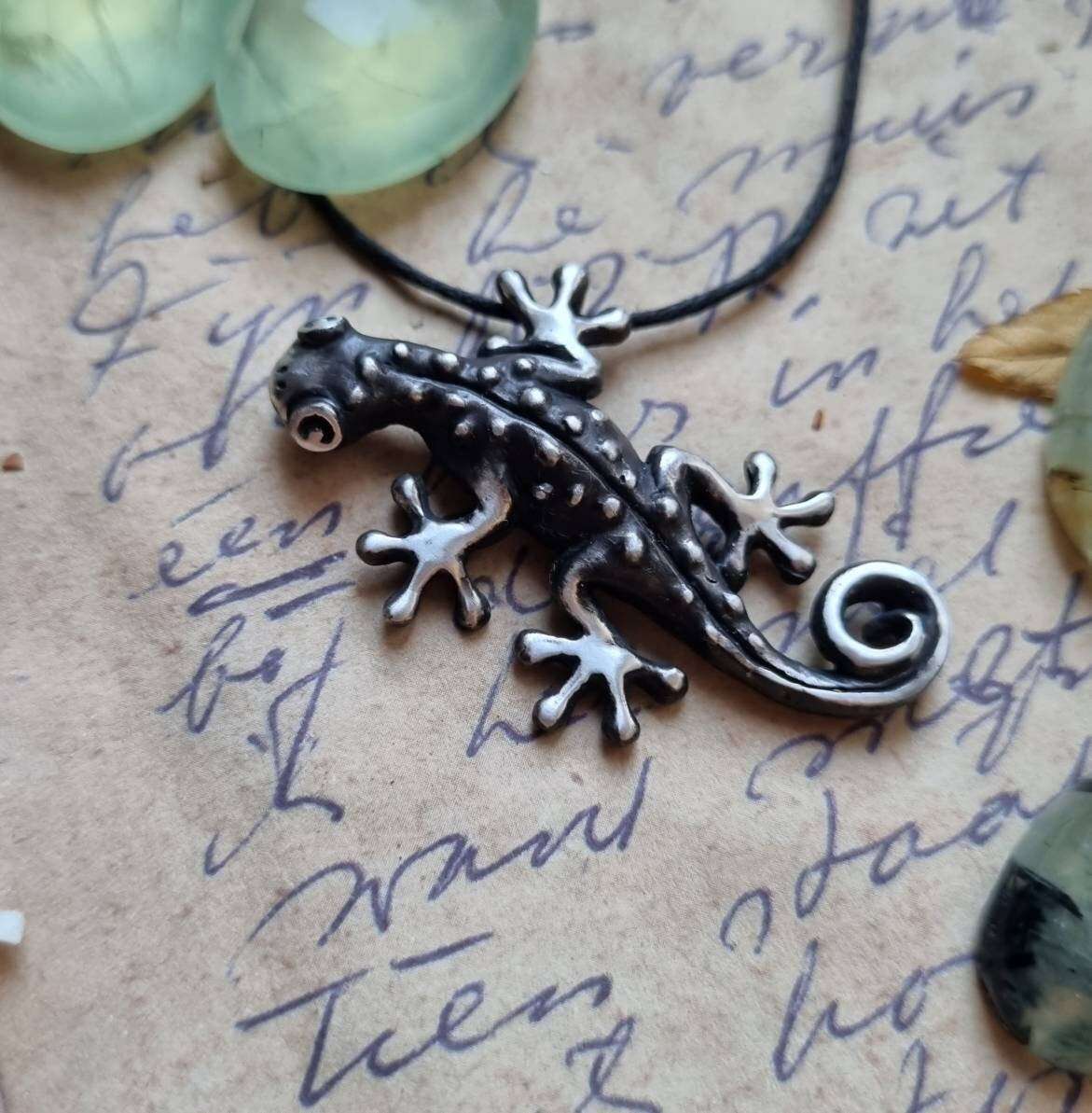 Sculptural silver gecko necklace photographed on handwritten paper with green gems in the background.
