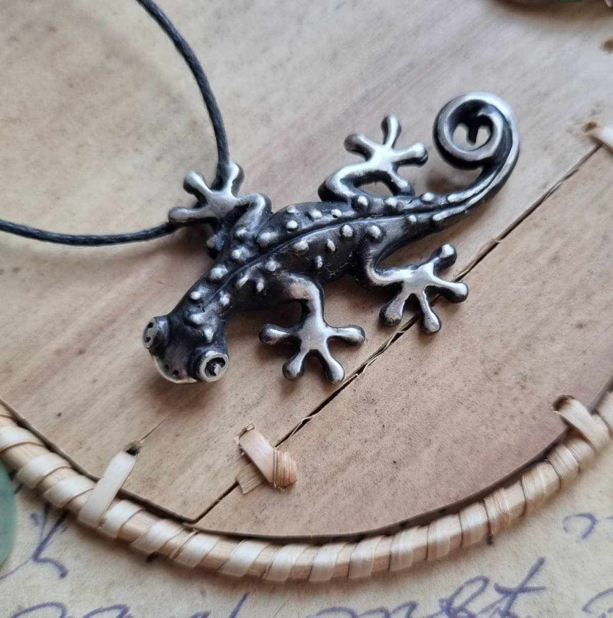 Handmade silver gecko pendant resting on a textured ceramic surface with rustic props.
