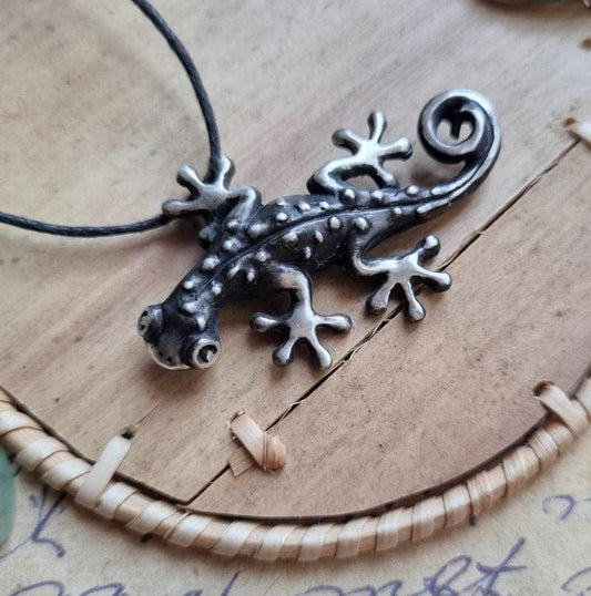 Handmade silver gecko pendant resting on a textured ceramic surface with rustic props.