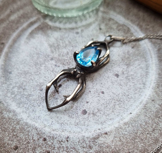 Silver Spider necklace with Swiss blue topaz on light ceramic plate — handcrafted sterling silver pendant with textured details and vivid blue gemstone.