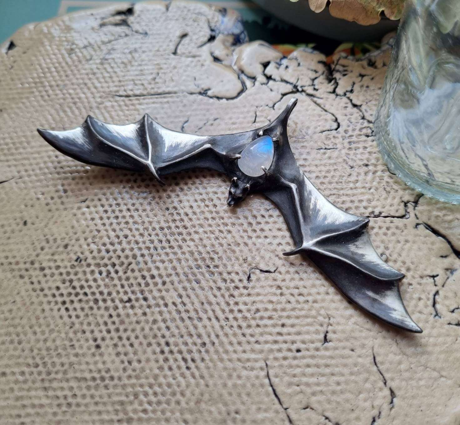 Handmade silver Bat pendant with rainbow moonstone, resting on natural textured background.