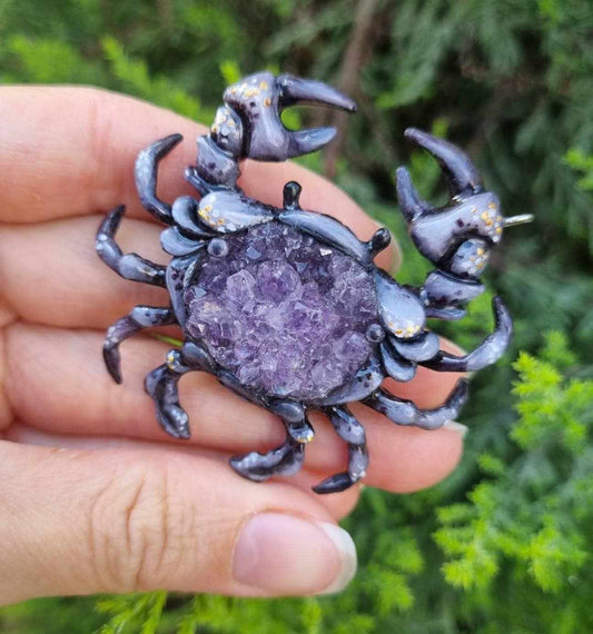 Handmade Crab necklace with amethyst druzy, held in hand against green foliage.