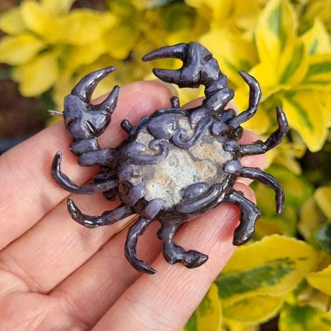 Back view of crab pendant showing painted details and structure, styled on natural foliage.