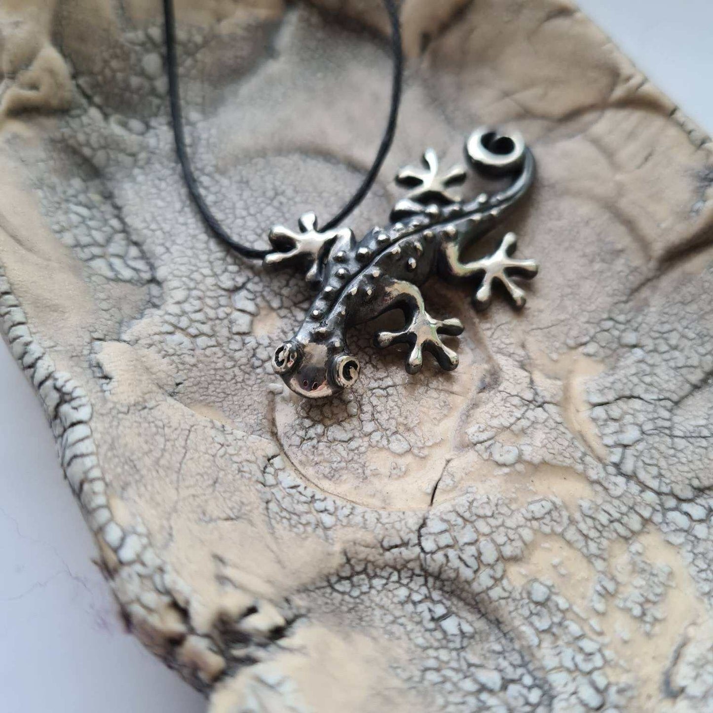 Gecko pendant in oxidized sterling silver lying on a textured ceramic plate, highlighting tactile texture.