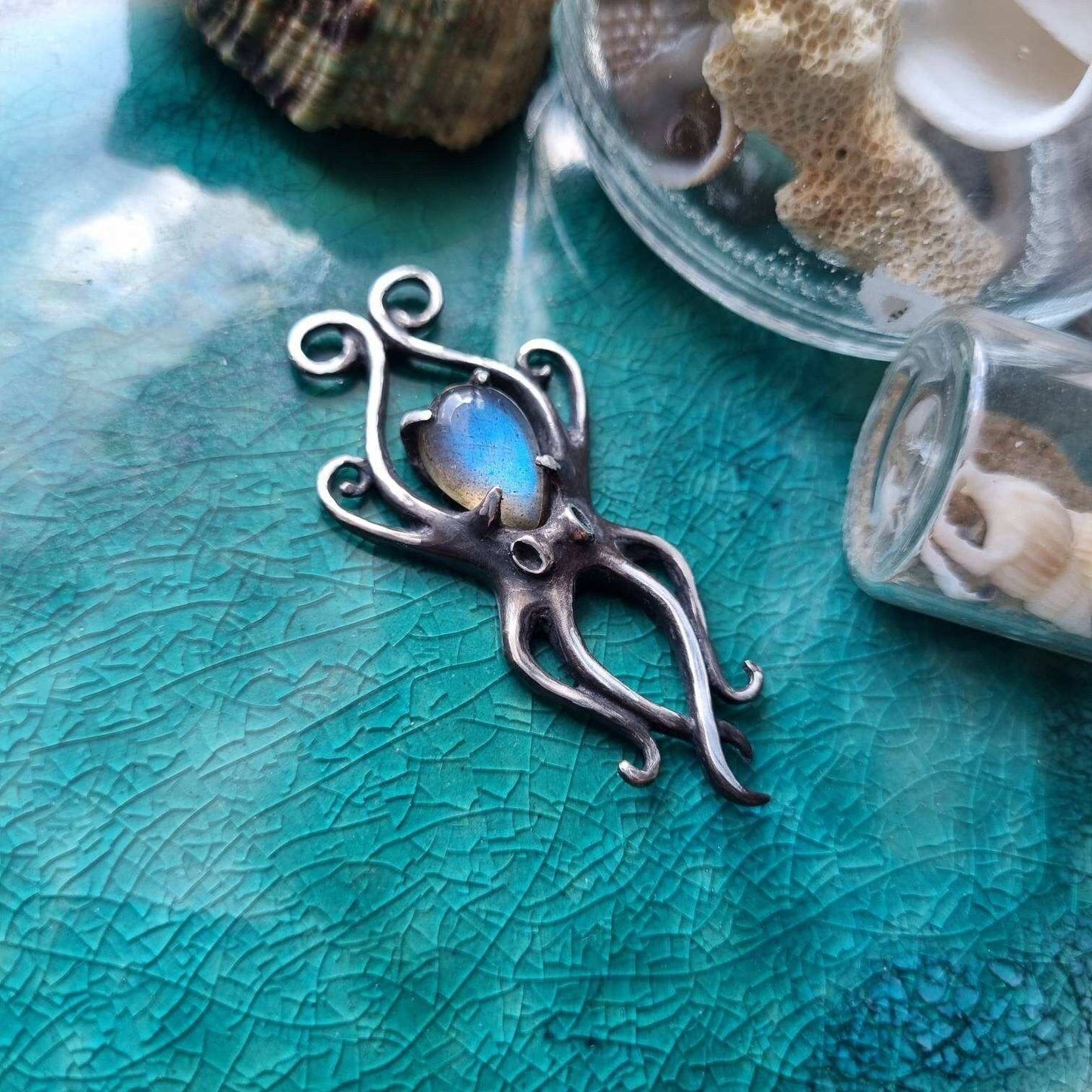 Sterling silver Octopus necklace with natural blue labradorite cabochon, photographed on textured teal surface.