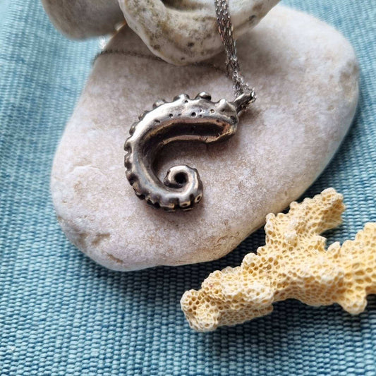 CloseClose-up of tentacle necklace resting on a blue background beside pale coral.-up of the octopus tentacle pendant with visible oxidized texture and sculptural detail.