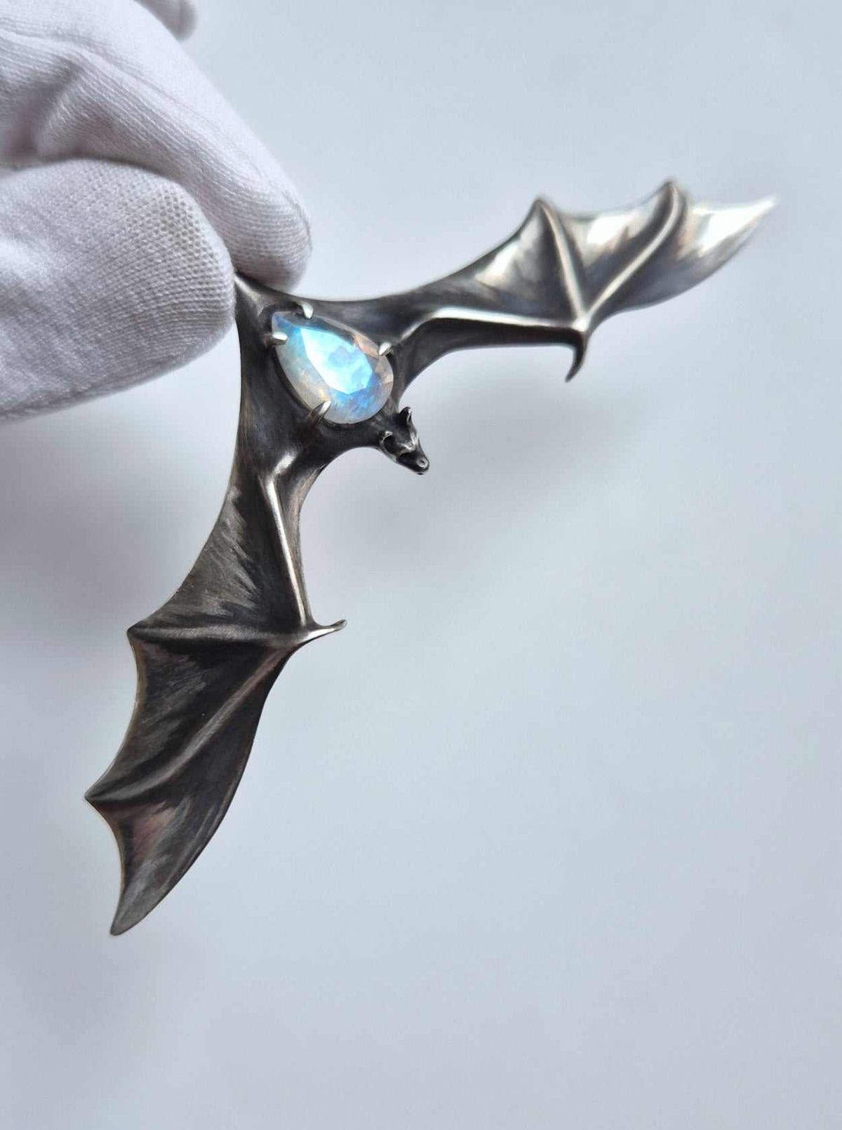 Silver bat pendant with blue-flash rainbow moonstone held by one wing in a white glove