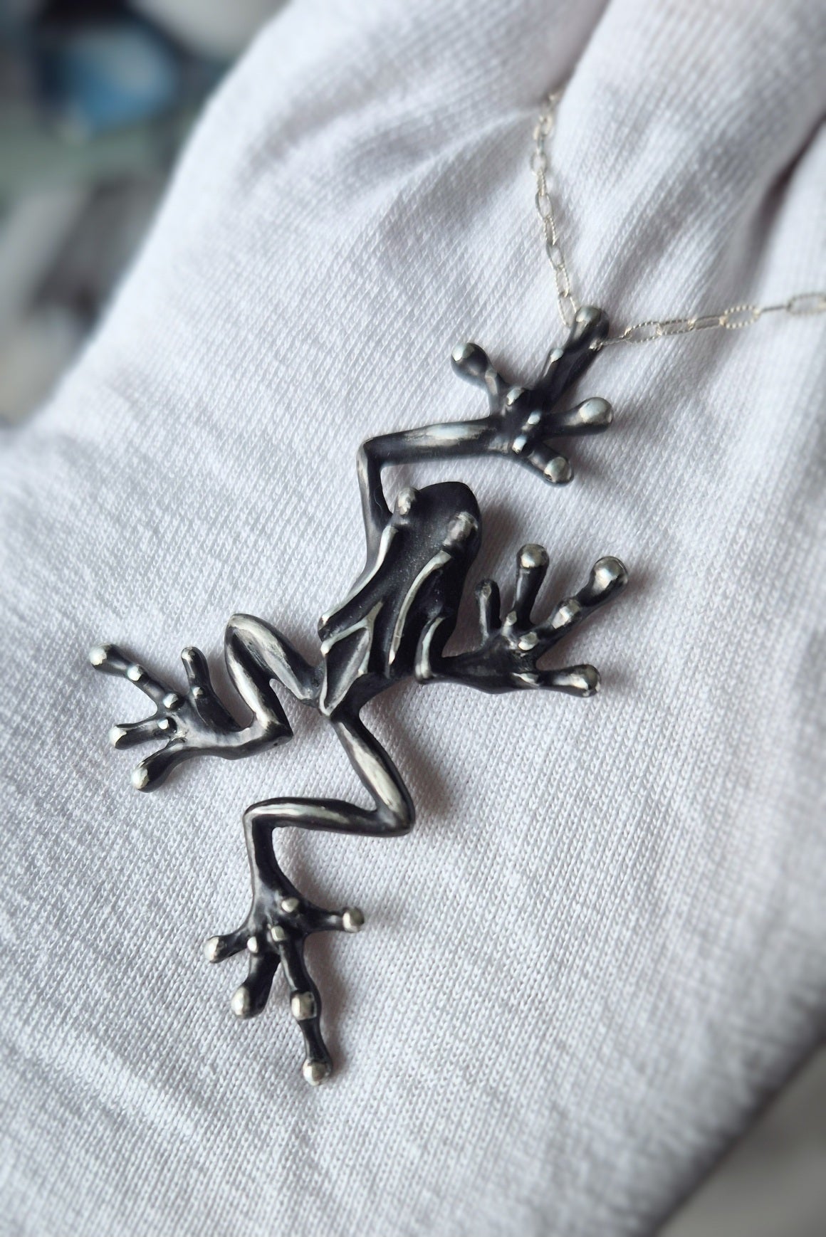 Angled close-up of a sculpted silver frog pendant displayed on white fabric