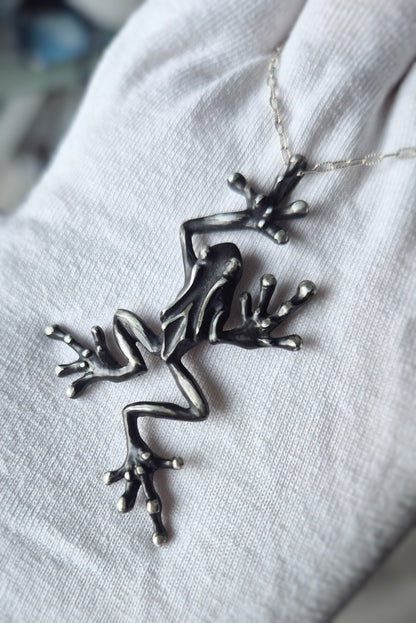 Angled close-up of a sculpted silver frog pendant displayed on white fabric