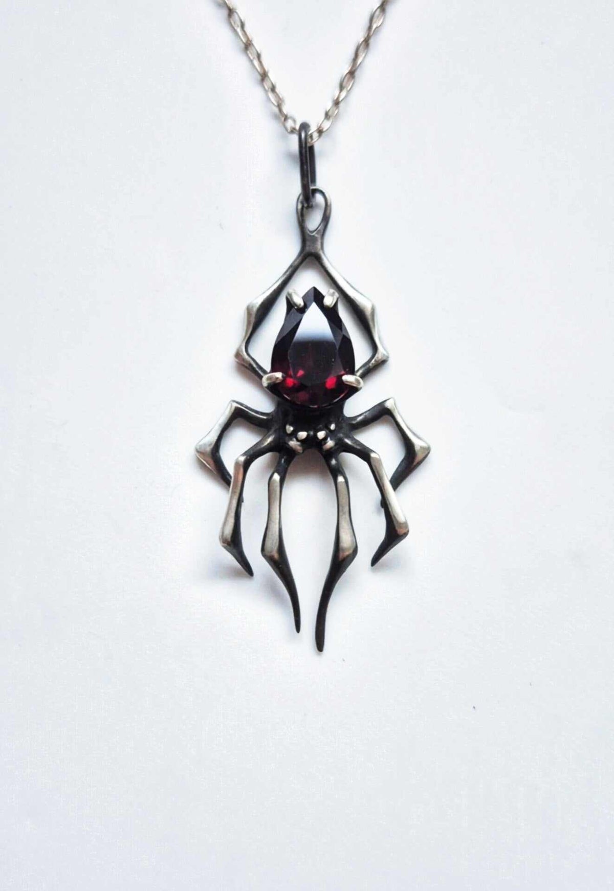 Front view of an oxidized sterling silver spider necklace with a pear-cut deep red garnet and chain on white