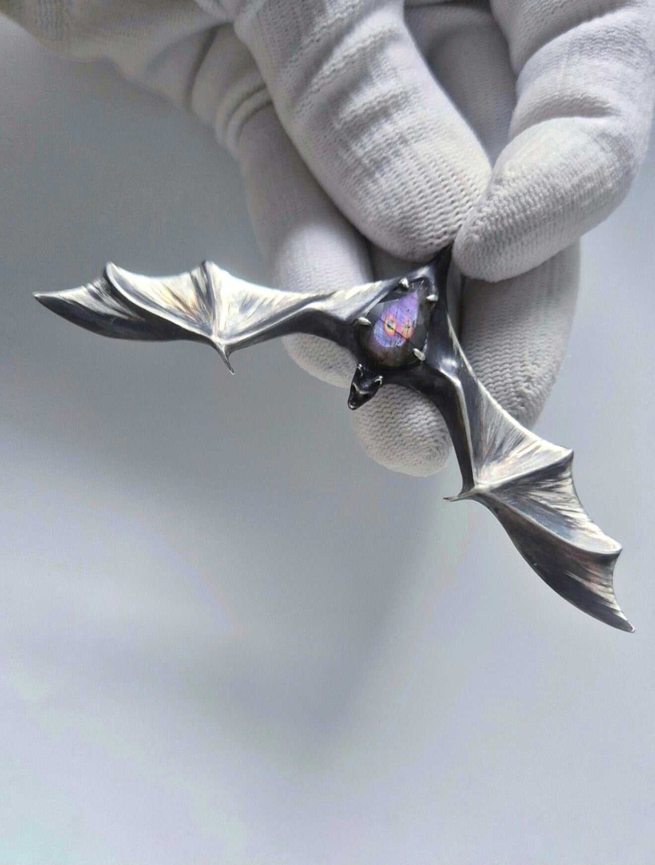 Oxidized silver bat pendant with purple-flash labradorite held by its bail in a white-gloved hand
