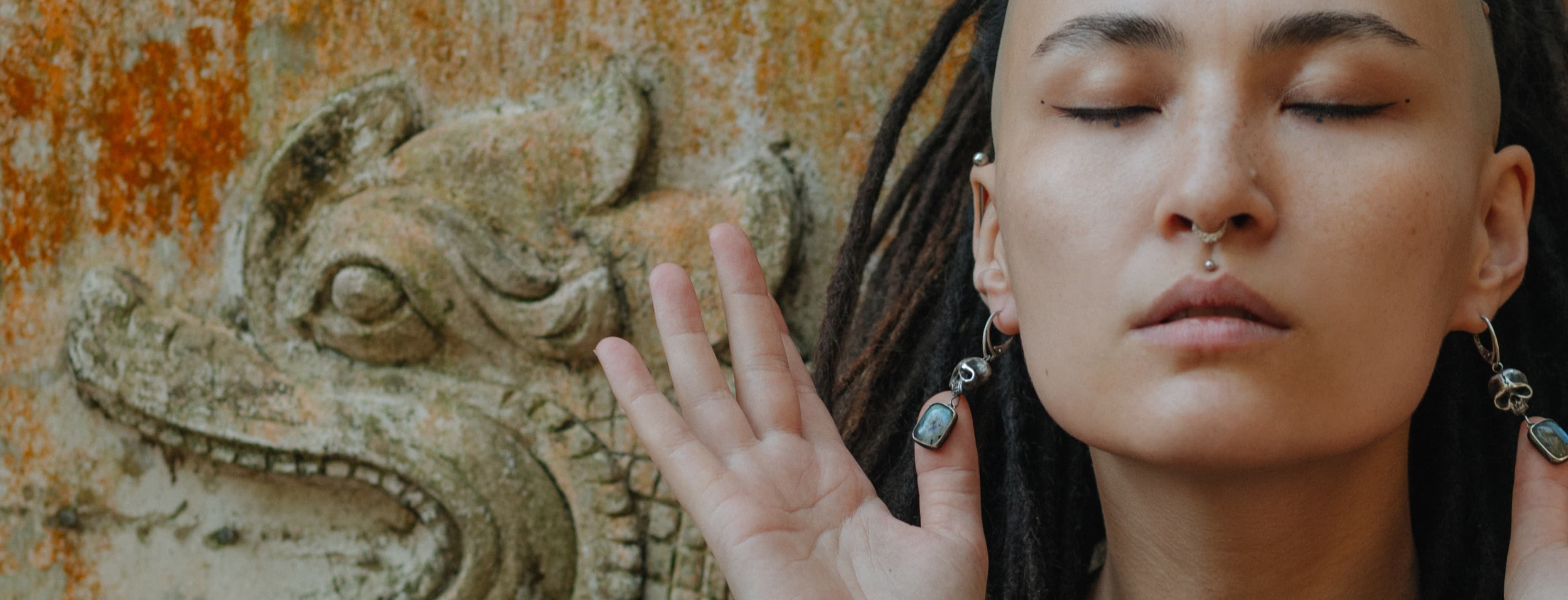 Person with dreadlocks and piercings wearing scull earrings in front of a decorative wall.