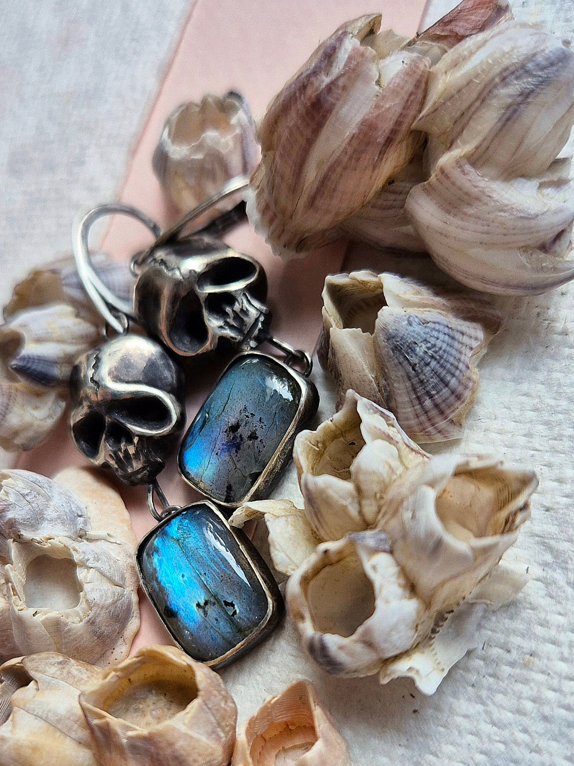 Close-up of handmade oxidised silver Skull earrings with blue labradorites, in a pile of barnacles and seashells