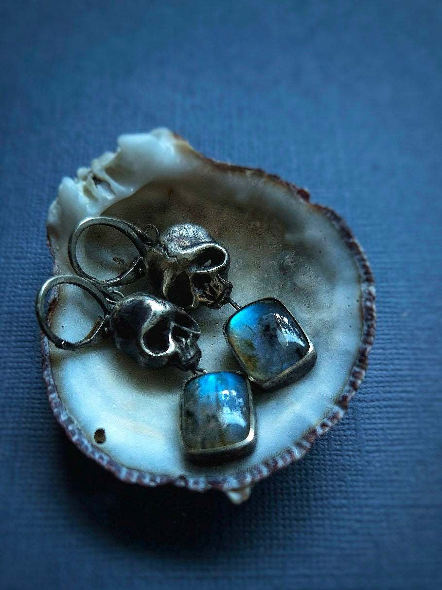 Close-up of handmade oxidised silver Skull earrings with blue labradorites in a seashell, in dim blue light