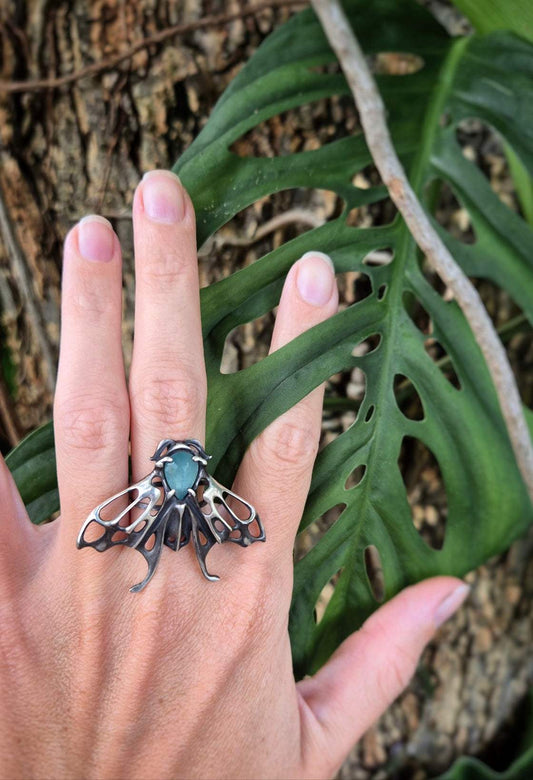 A front view of a Moth ring with a teal grandidierite gem, on a woman's hand with grean leaves and bark background