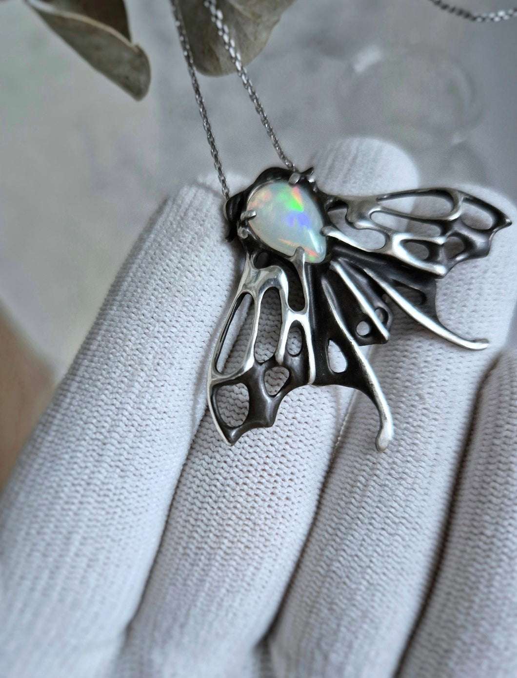 Macro shot of sculpted silver moth necklace and iridescent opal gem, displayed in glove for scale and craftsmanship.