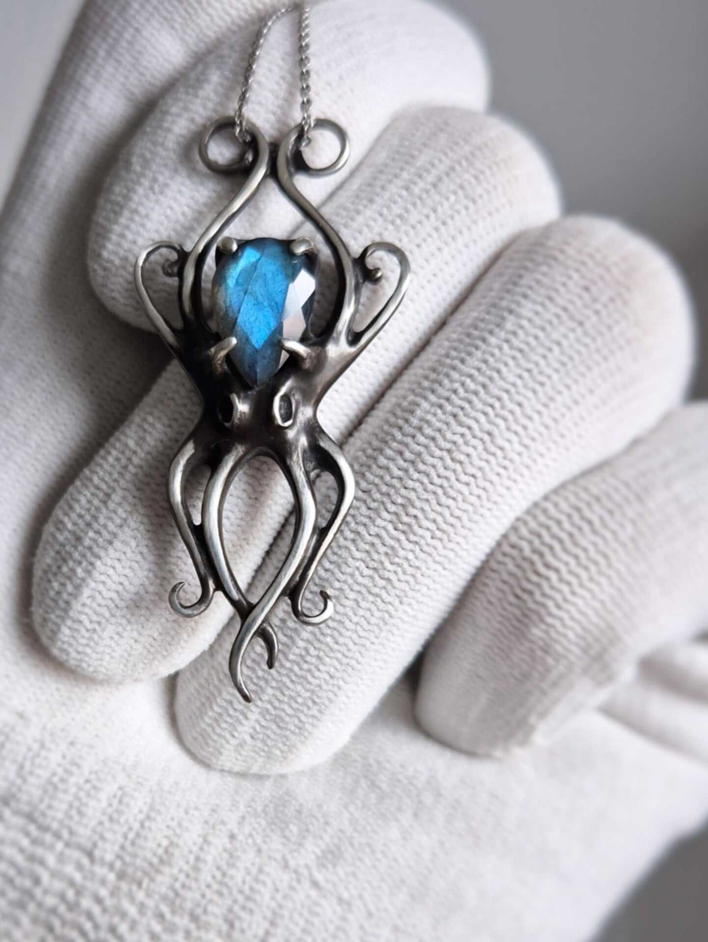 Angled macro of octopus pendant with glowing blue gemstone, sculpted tentacles visible, premium handcrafted silver jewelry image.