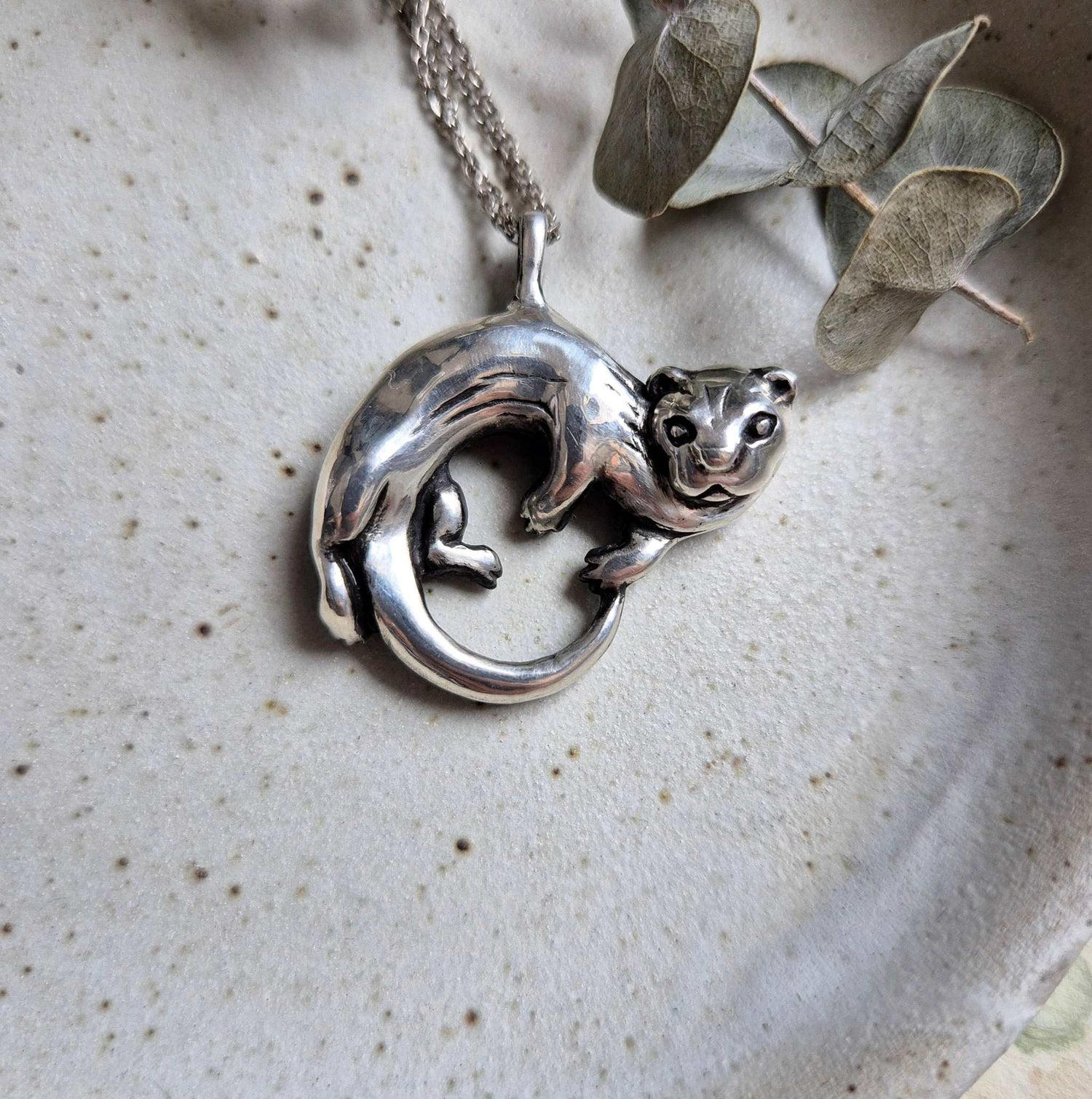 Close-up of the silver Otter necklace on a neutral ceramic background, highlighting the curved, sculptural shape of the pendant.