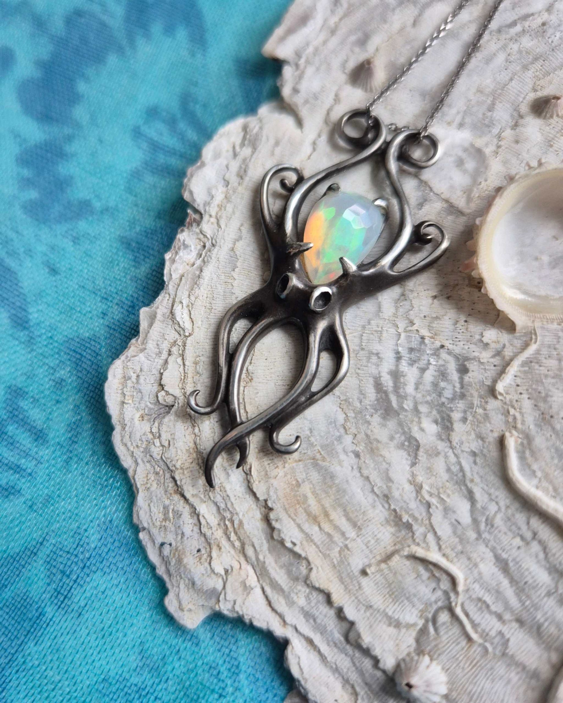 Handmade silver octopus pendant featuring an iridescent opal, photographed on a seashell surface