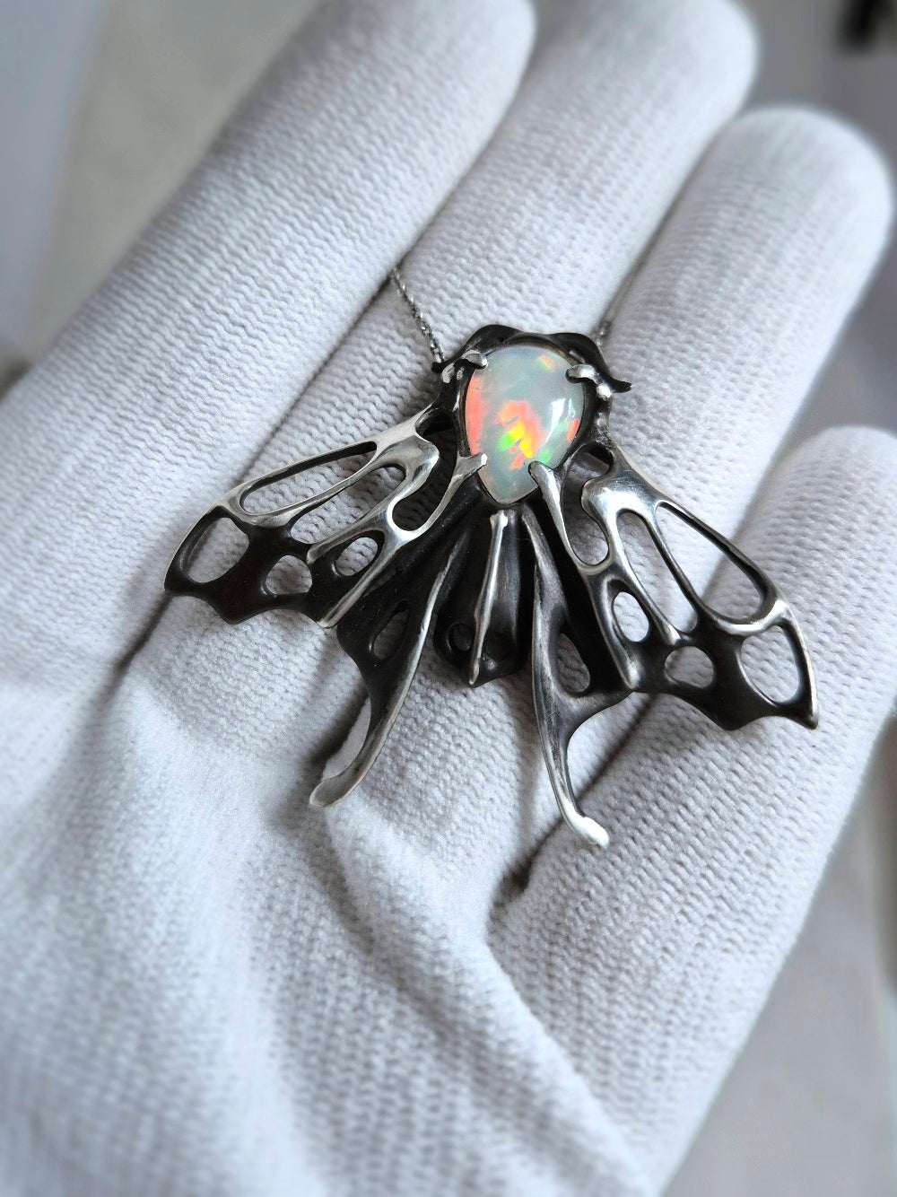 Handmade silver moth necklace with fiery opal gemstone held in white-gloved fingers, intricate wings fully spread.