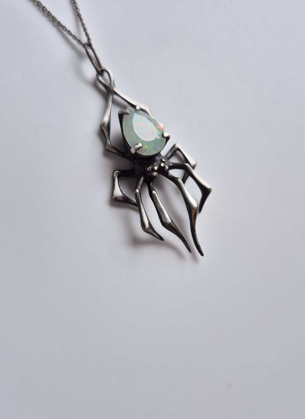 Artisan silver spider necklace with opal, tilted sideways on gradient background