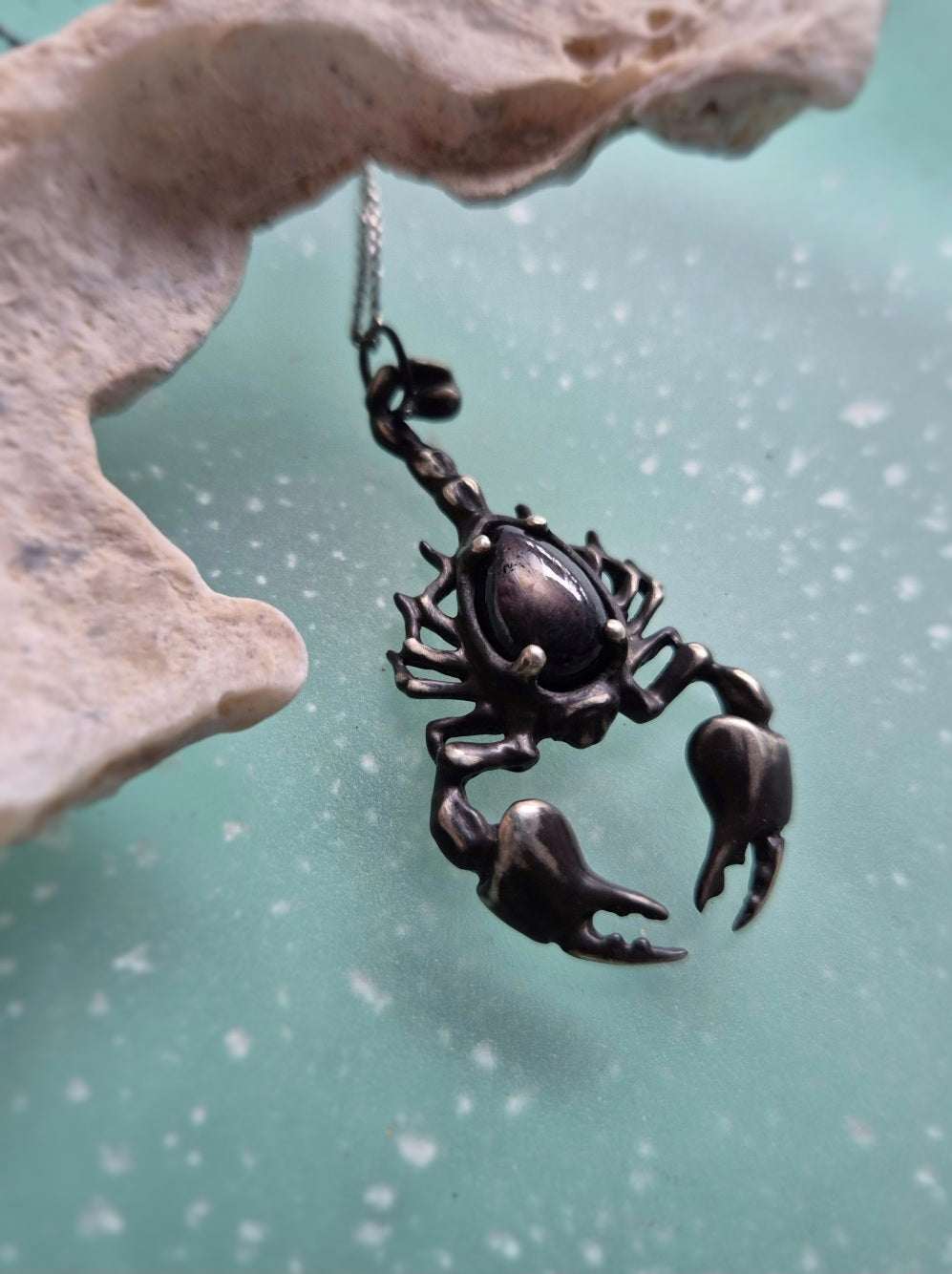 close-up of a silver Scorpion necklace with black gemstone on dark turquoise background