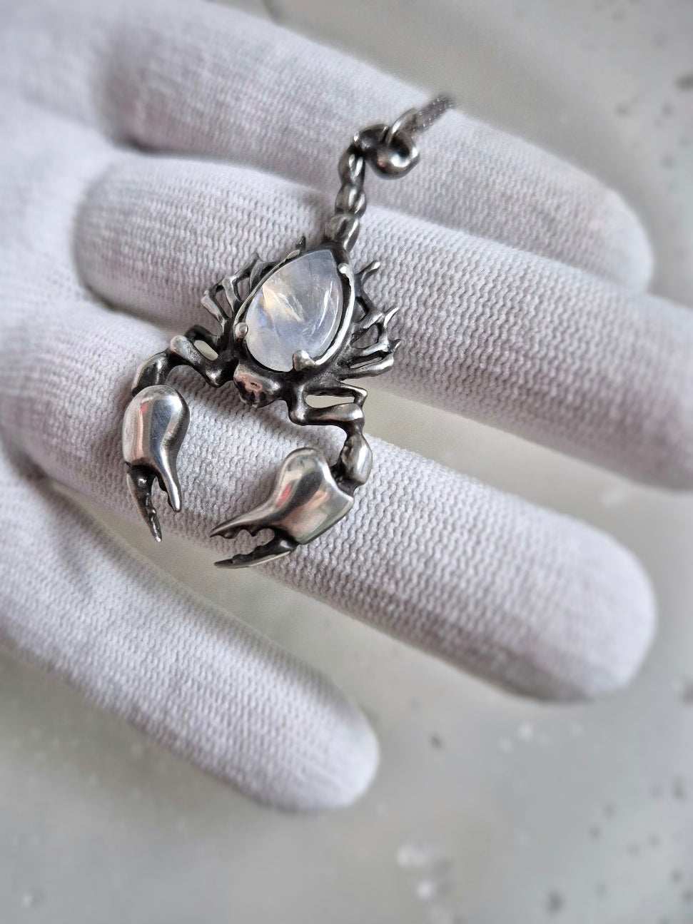 Handmade silver Scorpion necklace with luminous rainbow moonstone held in white-gloved fingers, detailed stinger visible.