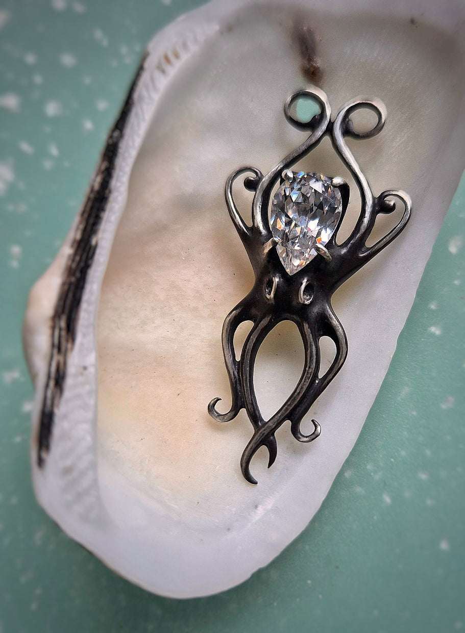 Octopus pendant in oxidized silver held in a white shell, showing sculptural tentacles and sparkling zircon.