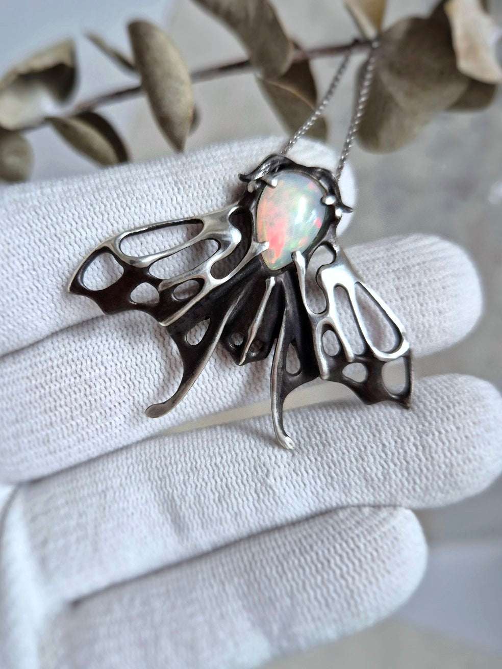 Close-up of sterling-silver moth pendant featuring oval opal cabochon, resting on gloved palm, detailed antennae visible.