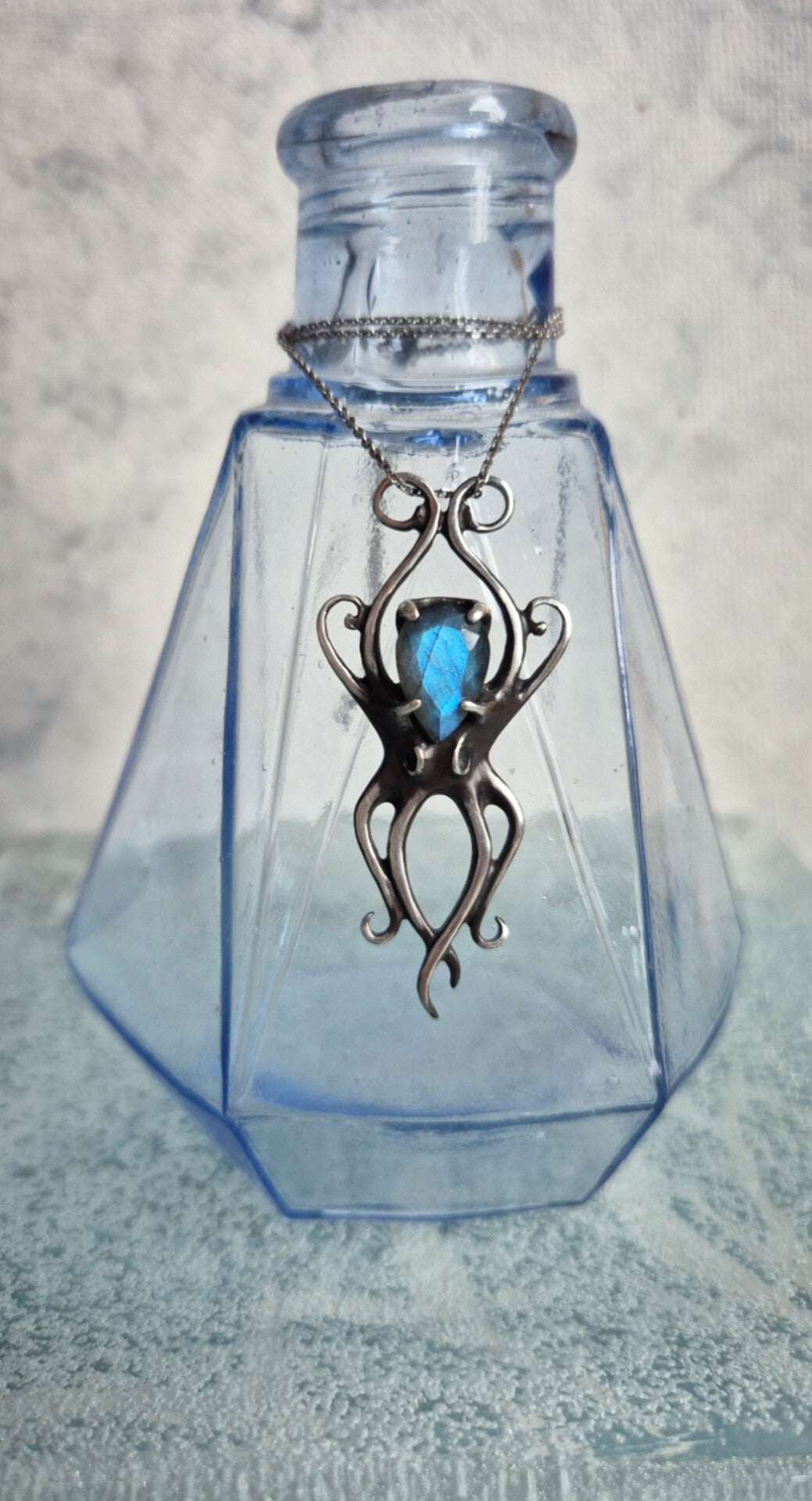Silver octopus necklace with vivid blue gemstone displayed inside clear blue glass bottle, artistic marine jewelry product shot.