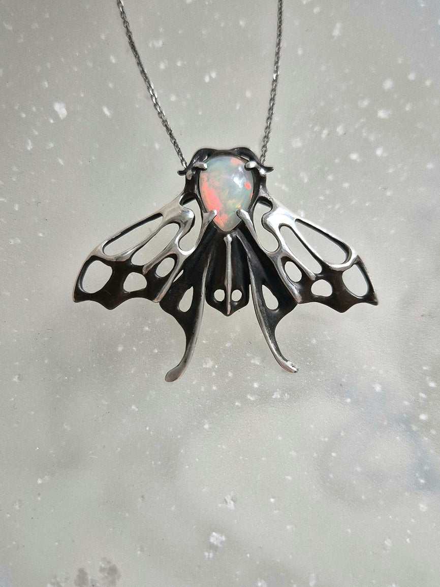 Silver moth pendant hanging from chain, opal stone shimmering, artisan insect jewelry on light marble surface.
