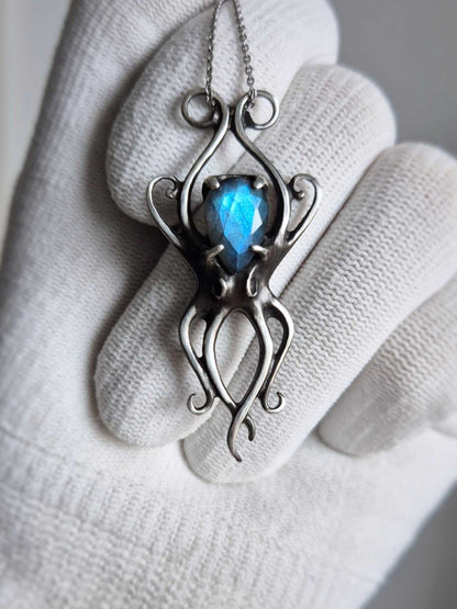 Close-up of handcrafted silver octopus pendant with electric-blue cabochon, held in white glove for detail view.