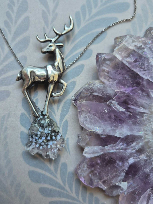 Handcrafted deer pendant with clear quartz crystal, shown beside amethyst cluster on soft blue paper