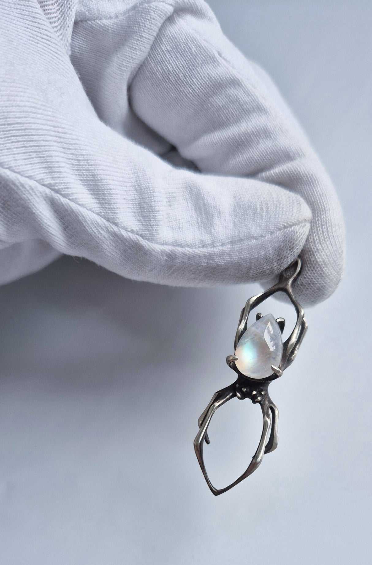 Full view of a silver spider pendant with pear-shaped rainbow moonstone held by its bail in a white glove