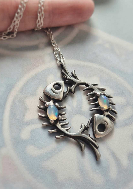 Sterling silver fish skeleton pendant with two Ethiopian opals held by its chain over pale patterned fabric