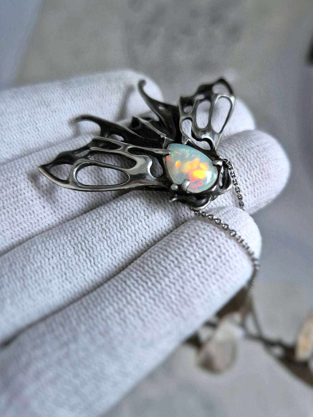 Side view of oxidized silver moth necklace with glowing opal center, photographed against neutral studio background.