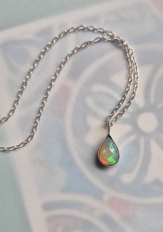 Silver opal pendant on chain, resting on a soft patterned background with subtle floral motif