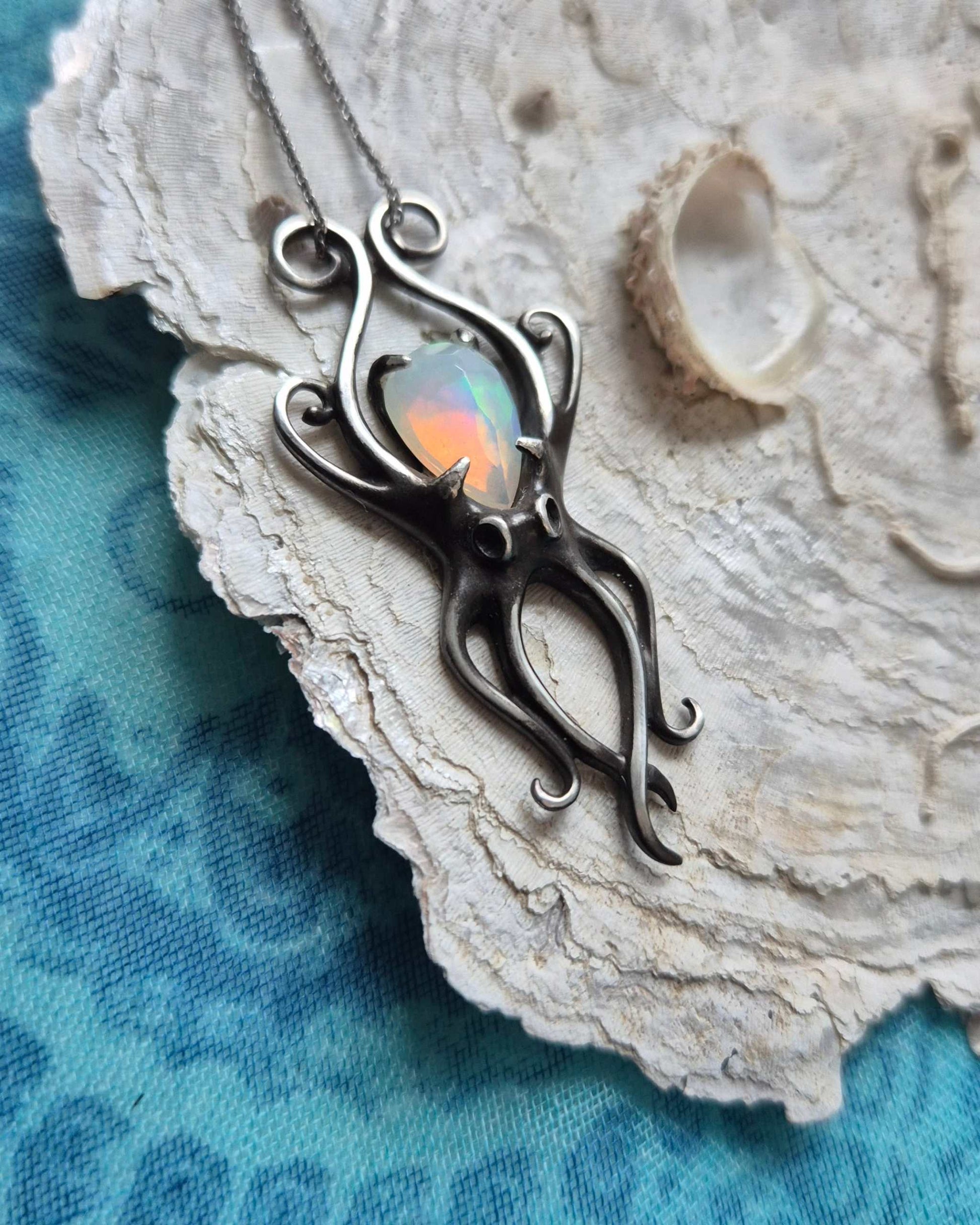 Silver octopus jewelry piece with glowing opal gemstone, arranged on a seashell over blue fabric