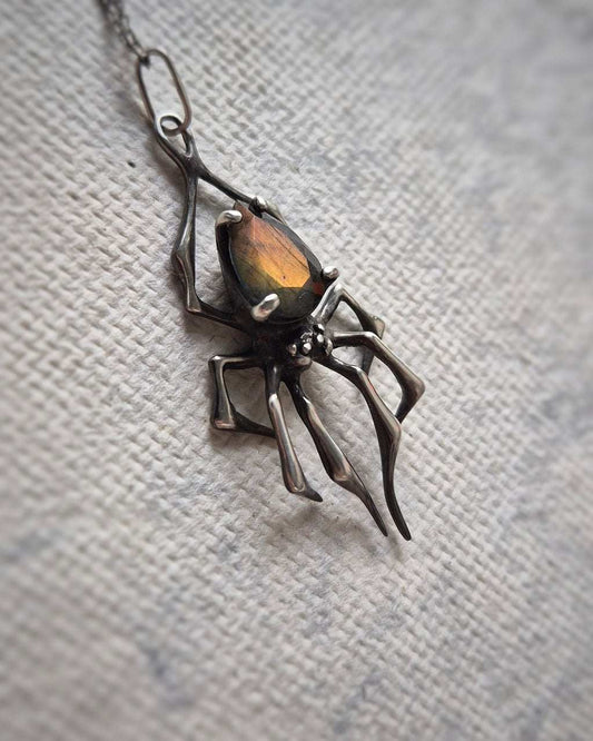 Close-up of a silver spider necklace with a glowing labradorite gemstone, lying on a light surface